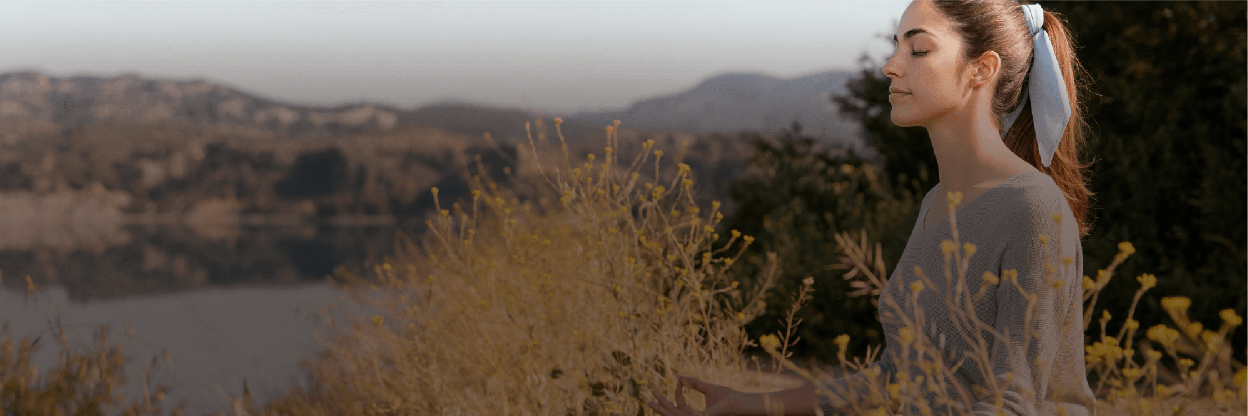 A woman sitting in stillness on a hilltop overlooking a lake at golden hour
